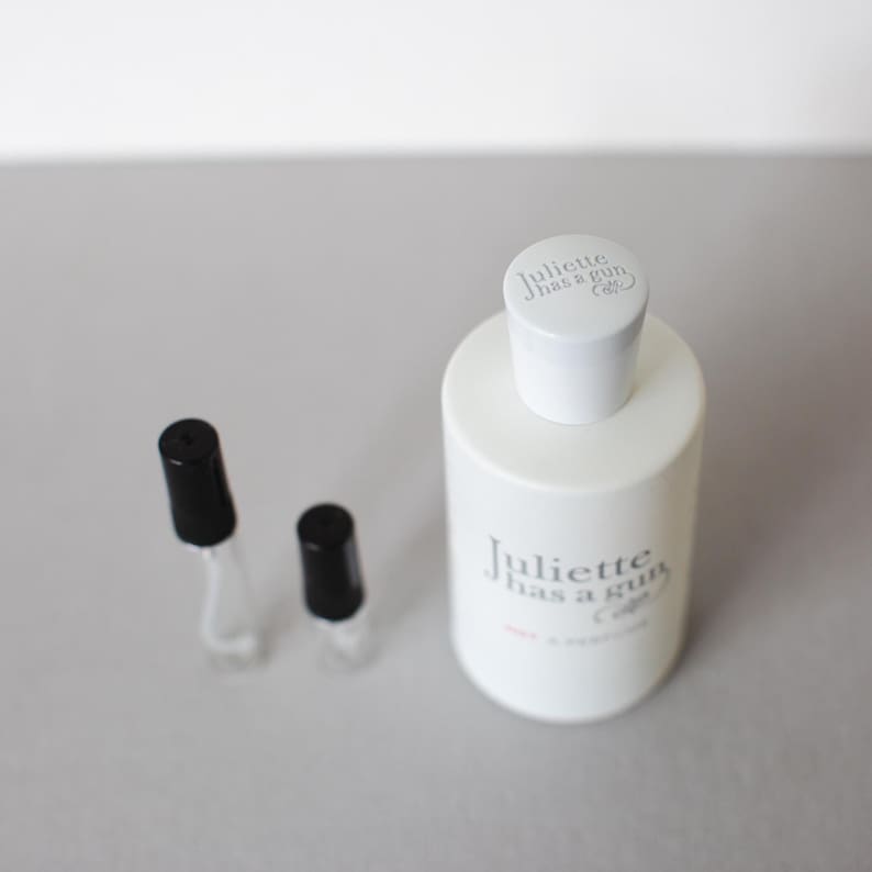 May include: A white perfume bottle with the text "Juliette has a gun" and two small sample vials with black caps. The bottle has a white cap with the brand name printed on it.