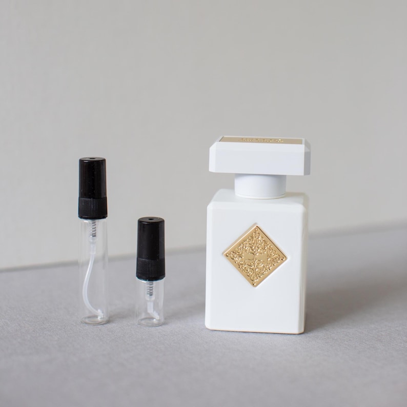 May include: A white perfume bottle with a gold emblem and a white cap, alongside two small glass vials with black caps. The perfume bottle is square-shaped and has a gold emblem on the front.