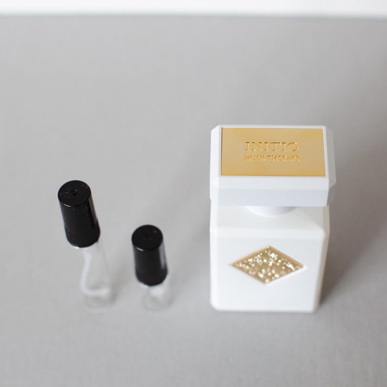 May include: A white perfume bottle with a gold top and a diamond-shaped glitter design. Two small, empty glass vials with black caps are next to the bottle. The bottle has the word "INITIO" printed on the top.