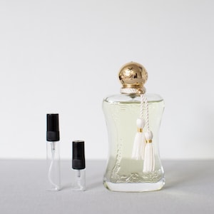 May include: A clear perfume bottle with a gold cap and white tassels, alongside two small sample vials with black caps. The perfume bottle has a decorative design and contains a light yellow liquid.