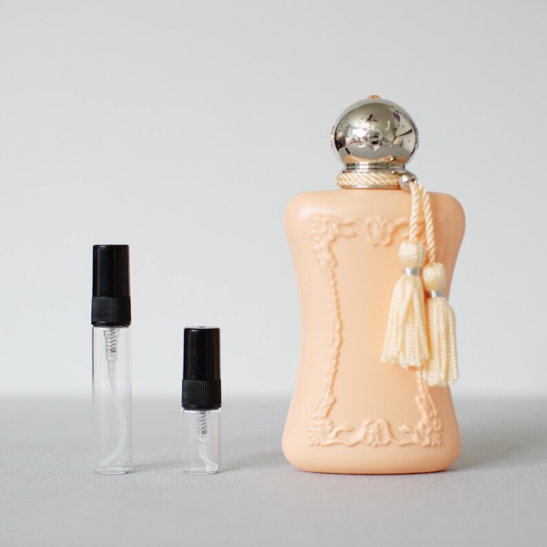 May include: A peach-coloured perfume bottle with a silver cap and two cream-coloured tassels, accompanied by two small, clear glass sample vials with black caps. The main bottle features an ornate, raised rectangular frame design on its front.
