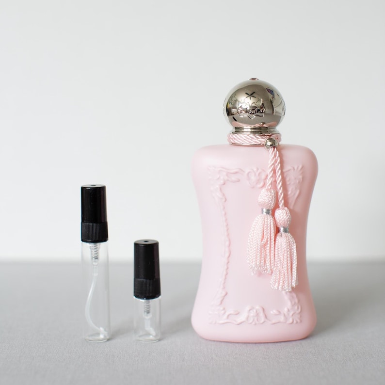 May include: A light pink perfume bottle with a silver cap and pink tassels. Two small, clear glass vials with black caps are next to the perfume bottle. The perfume bottle has a decorative floral design.