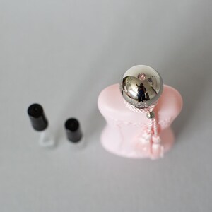May include: A silver-colored, spherical bottle stopper with a pink base and a tassel. Two black-capped bottles are in the background. The stopper reflects the surrounding environment. The overall theme is beauty and fragrance.