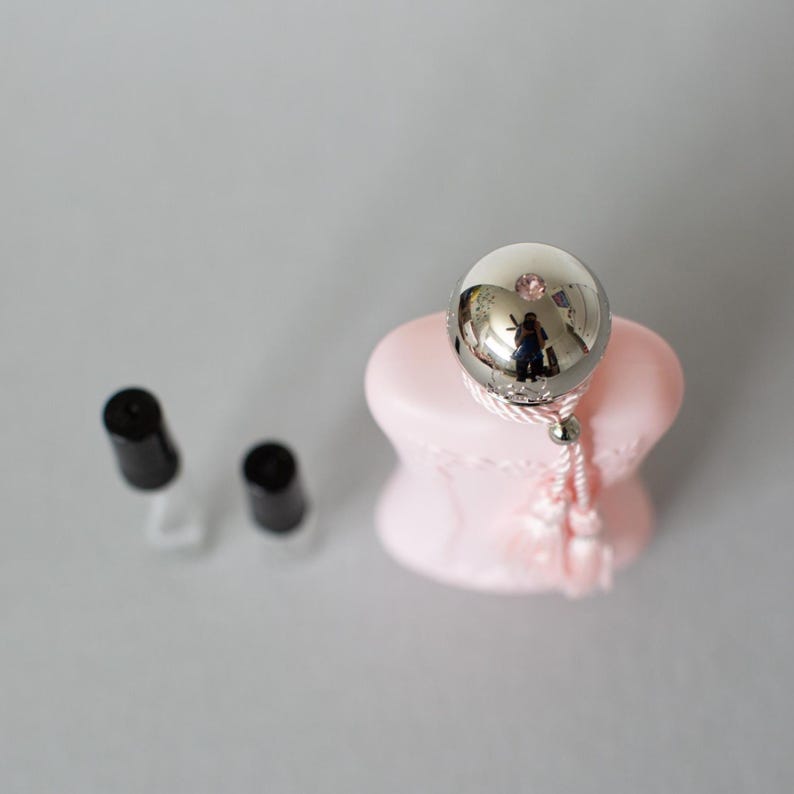 May include: A silver-colored, spherical bottle stopper with a pink base and a tassel. Two black-capped bottles are in the background. The stopper reflects the surrounding environment. The overall theme is beauty and fragrance.