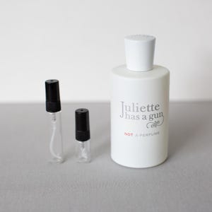 May include: A white perfume bottle with a white cap and the text "Juliette has a gun NOT A PERFUME". Two small, clear glass vials with black caps are next to the bottle.