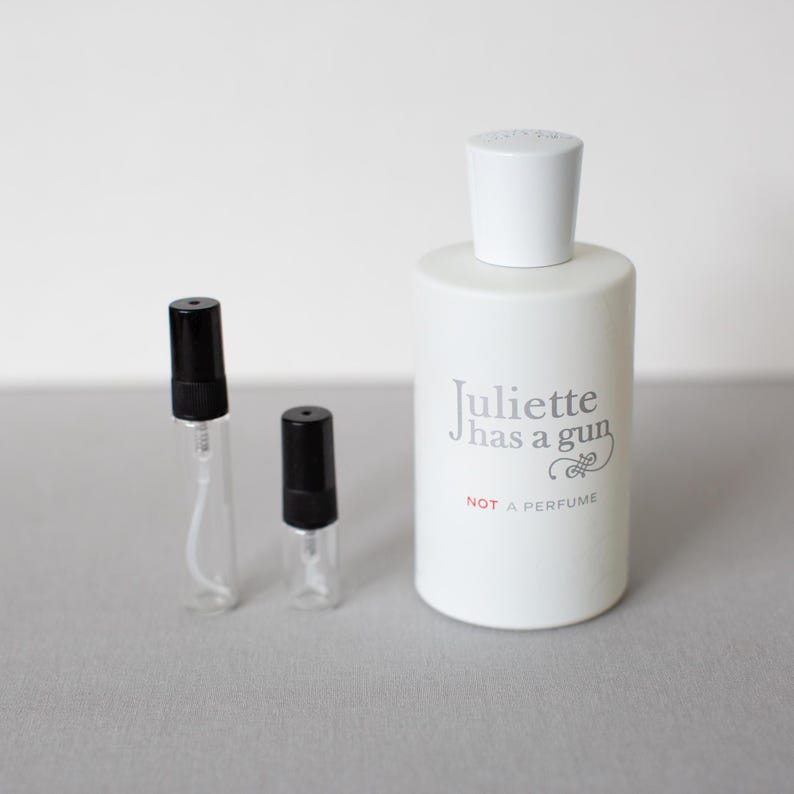 May include: A white perfume bottle with a white cap and the text "Juliette has a gun NOT A PERFUME". Two small, clear glass vials with black caps are next to the bottle.