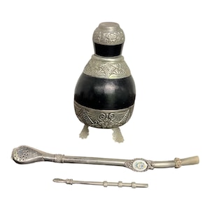 May include: A black gourd mate cup with silver-toned metal accents featuring floral engravings and three decorative feet, accompanied by a long metal bombilla straw with a perforated filter and a small, thin metal stirring tool with three rings.