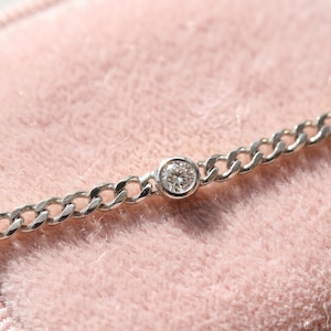 May include: A silver chain bracelet with a single, round, clear gemstone set in the center. The bracelet has a curb chain design and is displayed on a pink surface. The text "SHIVTARA JEWELS" is visible.