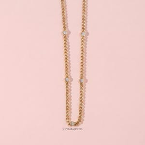 May include: A gold-colored chain necklace with five small, round, clear gemstones evenly spaced along its length. The necklace is displayed against a soft pink background. The brand name "SHIVTARA JEWELS" is printed at the bottom.