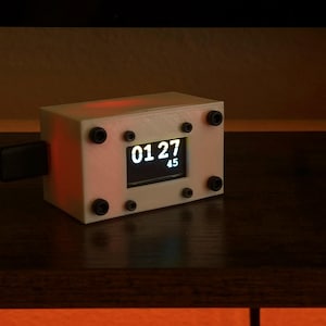May include: A small, white rectangular digital clock with a black display showing the time 01:27:45. The clock is secured with black screws and has a charging cable plugged in. The background is dark.