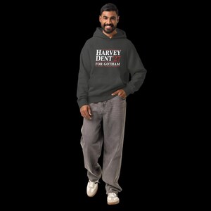 May include: A charcoal gray hoodie with the text "HARVEY DENT 27 FOR GOTHAM" in white and red. The person is wearing gray jeans and white sneakers. The hoodie has a hood and a front pocket.