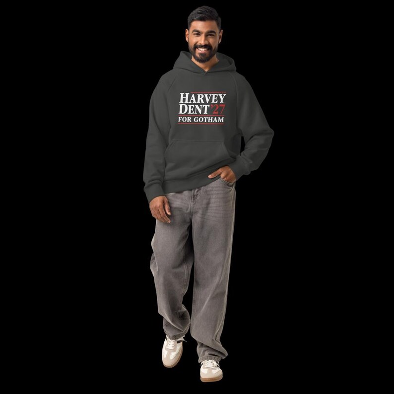 May include: A charcoal gray hoodie with the text "HARVEY DENT 27 FOR GOTHAM" in white and red. The person is wearing gray jeans and white sneakers. The hoodie has a hood and a front pocket.