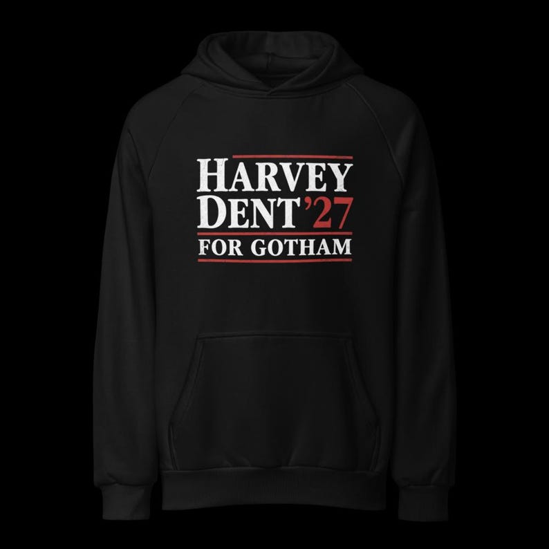May include: Black hooded sweatshirt with the text "HARVEY DENT '27 FOR GOTHAM" in white and red lettering. The sweatshirt has a front pocket and a hood. The design is centered on the chest.
