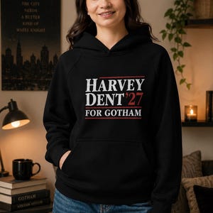 May include: Black hooded sweatshirt with the text "HARVEY DENT '27 FOR GOTHAM" in white and red lettering. The sweatshirt has a front pocket and is worn with blue jeans. A poster with the word "GOTHAM" is visible in the background.