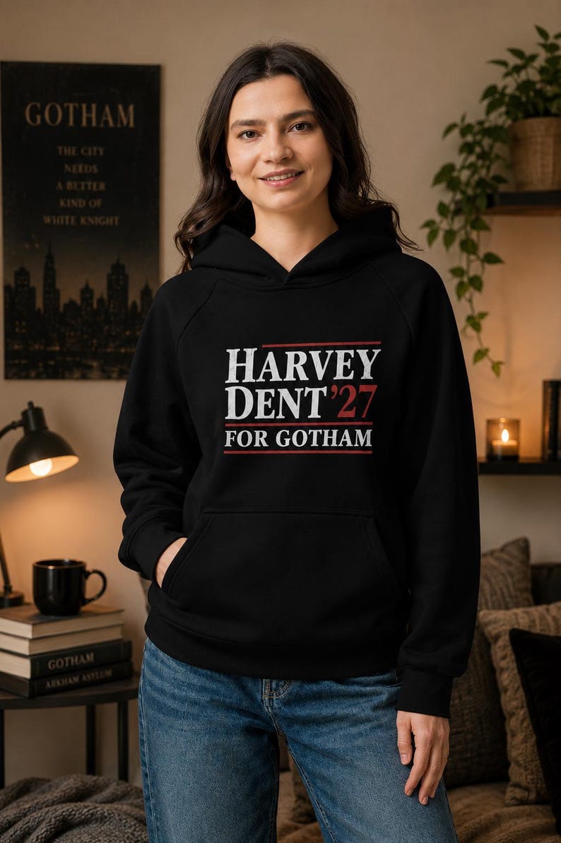 May include: Black hooded sweatshirt with the text "HARVEY DENT '27 FOR GOTHAM" in white and red lettering. The sweatshirt has a front pocket and is worn with blue jeans. A poster with the word "GOTHAM" is visible in the background.