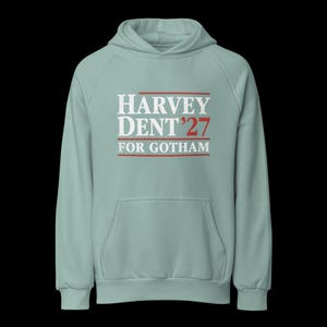 May include: A light green hoodie with a front pocket and a hood. The front of the hoodie features the text "HARVEY DENT '27 FOR GOTHAM" in white and red lettering. The hoodie is made of a soft material.