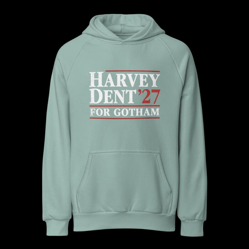 May include: A light green hoodie with a front pocket and a hood. The front of the hoodie features the text "HARVEY DENT '27 FOR GOTHAM" in white and red lettering. The hoodie is made of a soft material.