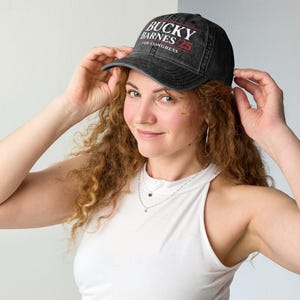 Könnte beinhalten: Eine schwarze Baseballkappe mit dem Text "BUCKY BARNES FOR CONGRESS 25" in Weiß und Rot. Die Kappe wird von einer Person mit langen, lockigen roten Haaren und einem weißen Oberteil getragen.