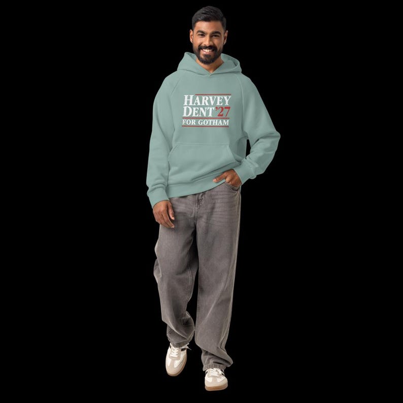 May include: A light blue hoodie with the text "HARVEY DENT 27 FOR GOTHAM" in white and red lettering. The person is wearing gray jeans and white sneakers. The hoodie has a front pocket and a hood.