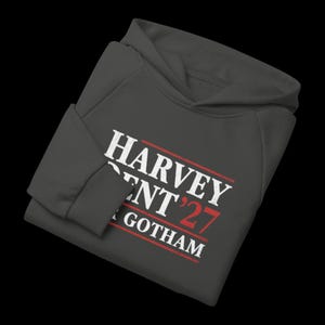May include: Dark gray hoodie with the text "HARVEY DENT '27 GOTHAM" in white and red. The hoodie has a hood and long sleeves. The text is in a bold, sans-serif font.