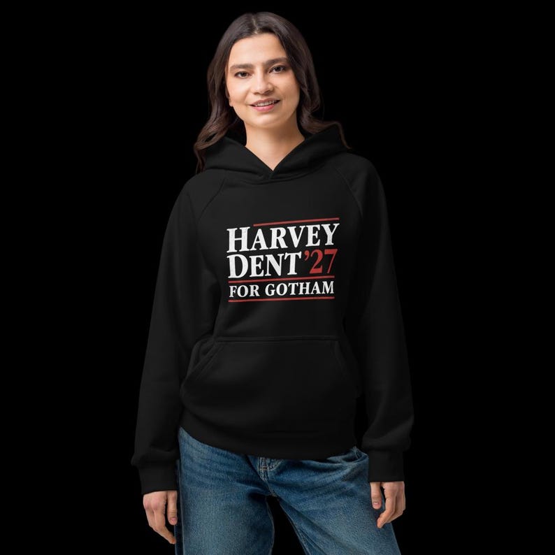 May include: Black hoodie with the text "HARVEY DENT '27 FOR GOTHAM" in white and red lettering. The hoodie has a front pocket and a hood. The person is wearing blue jeans.