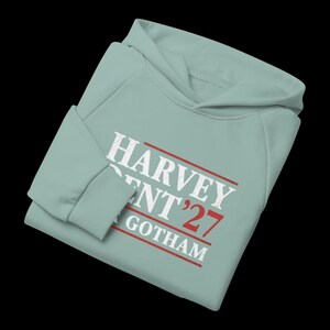 May include: A light blue hoodie with the text "HARVEY DENT '27 GOTHAM" in white and red. The hoodie has a hood and long sleeves. The text is in a bold, sans-serif font.