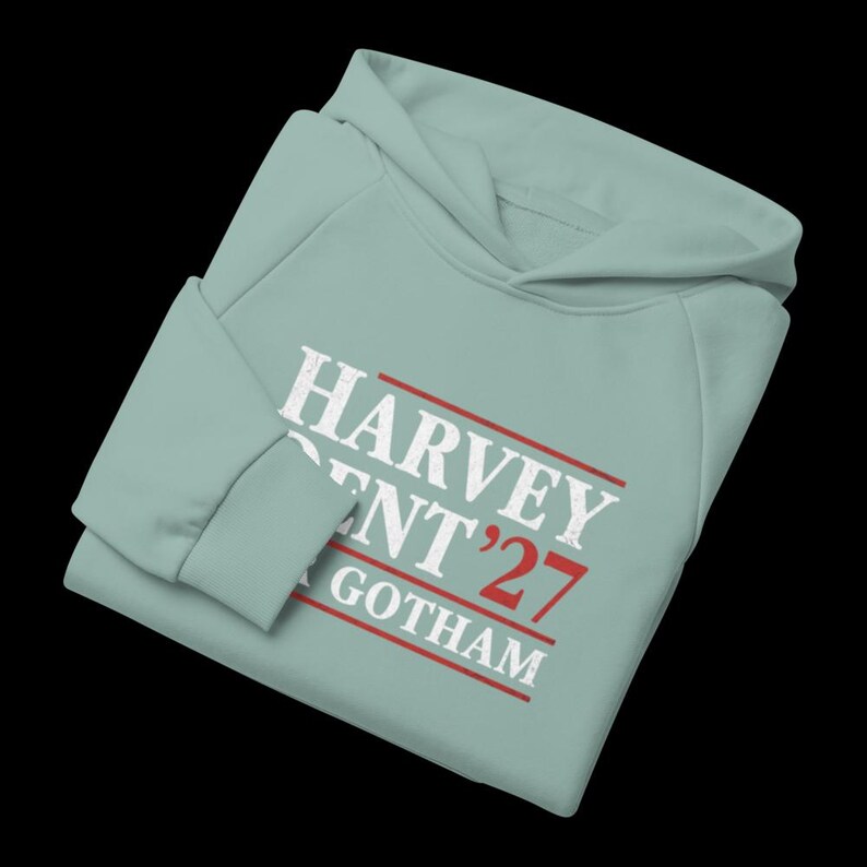 May include: A light blue hoodie with the text "HARVEY DENT '27 GOTHAM" in white and red. The hoodie has a hood and long sleeves. The text is in a bold, sans-serif font.