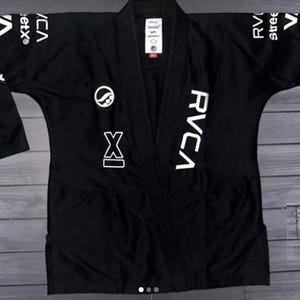 Op de afbeelding: Een zwart Braziliaans Jiu-Jitsu gi-jack met wit RVCA-logo op de linkerborst en mouwen. Het kledingstuk is voorzien van een cirkelvormig logo op de rechterborst, een Romeins cijfer XI-afbeelding en een wit label in de nek met zwarte tekst.