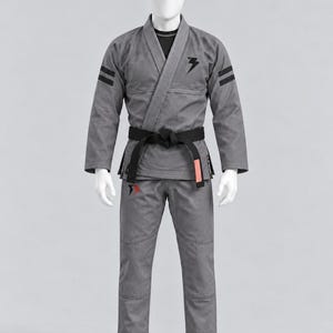 May include: A grey Brazilian Jiu-Jitsu gi featuring a black lightning bolt logo on the chest, two black stripes on each sleeve, and a black belt tied at the waist. The pants include a small red and black logo detail on the right thigh area.