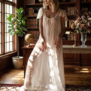 Puede incluir: Un camisón transparente de color marfil con mangas cortas y escote anudado. El camisón es largo hasta el suelo y fluido. La imagen está ambientada en una biblioteca con una gran ventana, estanterías y un escritorio. El tema general es lencería.