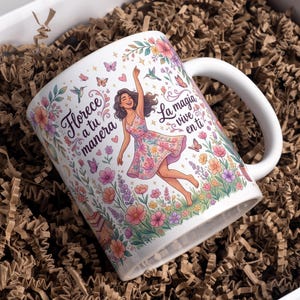 May include: A white ceramic mug featuring a colorful illustration of a person in a floral dress surrounded by flowers, butterflies, and hummingbirds. The text reads "Florece a tu manera" and "La magia vive en ti" in a black cursive script.