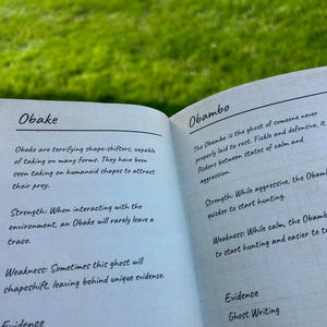 Pode incluir: Livro aberto com texto sobre Obake e Obambo, dois tipos de fantasmas. A descrição de Obake detalha suas habilidades de mudança de forma e fraquezas. A descrição de Obambo explica sua natureza volúvel e comportamentos de caça. Fundo de grama verde desfocada.