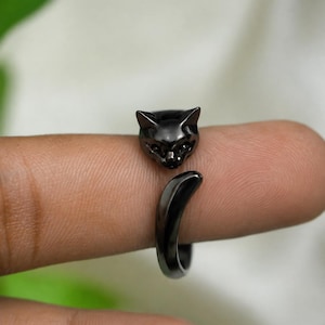 May include: A black metal ring featuring a cat head design at one end and a curved tail shape at the other. The ring has a polished, dark finish and an open band structure that wraps around the finger, showcasing detailed feline facial features.