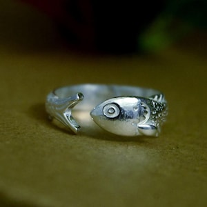 May include: A silver-toned open ring featuring a fish design where the head and tail ends meet. The fish has a detailed eye, textured scales along the band, and a fin. The ring is adjustable, with a gap between the head and the tail of the fish.