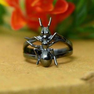 May include: A dark metallic ring featuring a sculpted ant design on the front. The insect figure is centered on the band, showcasing detailed legs, antennae, and a segmented body. The ring has a smooth, polished finish with a deep charcoal gray tone.