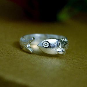 Peut inclure: Une bague ouverte argentée représentant un poisson. L'anneau est formé par le corps d'un poisson avec une tête détaillée à une extrémité et une nageoire caudale à l'autre, créant un espace ajustable. La surface présente un fini métallique poli.