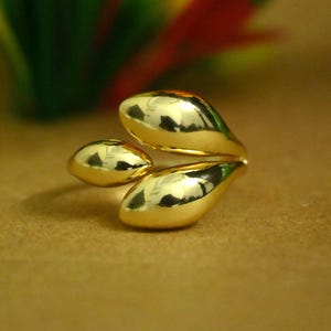 May include: A polished gold ring featuring three smooth, teardrop-shaped elements arranged in a cluster. The metal surface is highly reflective, showing highlights and shadows that emphasize the rounded, organic forms of the design.