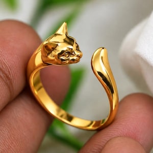 以下が含まれることがあります： 片方の端に彫刻された猫の頭、もう片方の端に滑らかで先細りの尻尾をあしらった、ゴールドトーンのオープンリング。バンドは磨き上げられて光沢があり、バイパススタイルで指を包み込む連続した金属面が特徴です。
