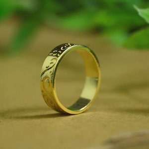 Pode incluir: Um anel de ouro com um design gravado. O anel tem uma banda com um padrão repetido de redemoinhos e curvas. O anel é feito de um material dourado brilhante e está sobre um fundo neutro.