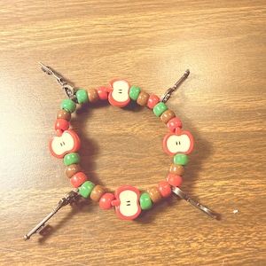Puede incluir: Una pulsera de cuentas con cuentas rojas en forma de manzana con detalles negros, intercaladas con cuentas verdes y marrones. La pulsera está conectada con cierres plateados. La pulsera está sobre una superficie de madera.