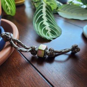 Könnte beinhalten: Ein beiges und schwarz gesprenkeltes Paracord-Lanyard mit einer zentralen, sechseckigen Metallmutter in Metallic-Optik. Die Schnur ist auf beiden Seiten der Mutter mit zwei Knoten versehen, mit einer Schlaufe an einem Ende und einem Knoten am anderen.