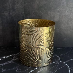 May include: A cylindrical gold-colored metal container featuring a repeating embossed pattern of fan-shaped leaves. The surface has a metallic sheen with visible vertical striations throughout the texture, creating a consistent decorative design.