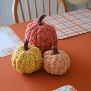 May include: Three plush, crocheted pumpkins in varying sizes and colors. The largest is a deep rust orange, the medium is a mustard yellow, and the smallest is a soft peach. Each pumpkin features a dark brown, textured stem on top.