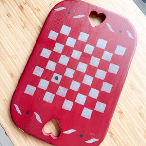May include: A rectangular red wooden serving tray with rounded corners, featuring a central silver checkerboard pattern. The tray includes two heart-shaped cutouts at the top and bottom, accented with small silver leaf motifs and dark blue dots.