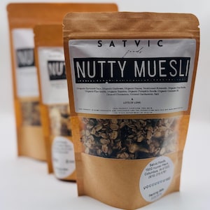 May include: Three brown paper bags of "Nutty Muesli" are displayed. The front bag has a clear window showing the granola inside. The label reads "Satvic Foods" and lists ingredients like oats, cashews, and dates. The text "Lots of Love" is also visible.