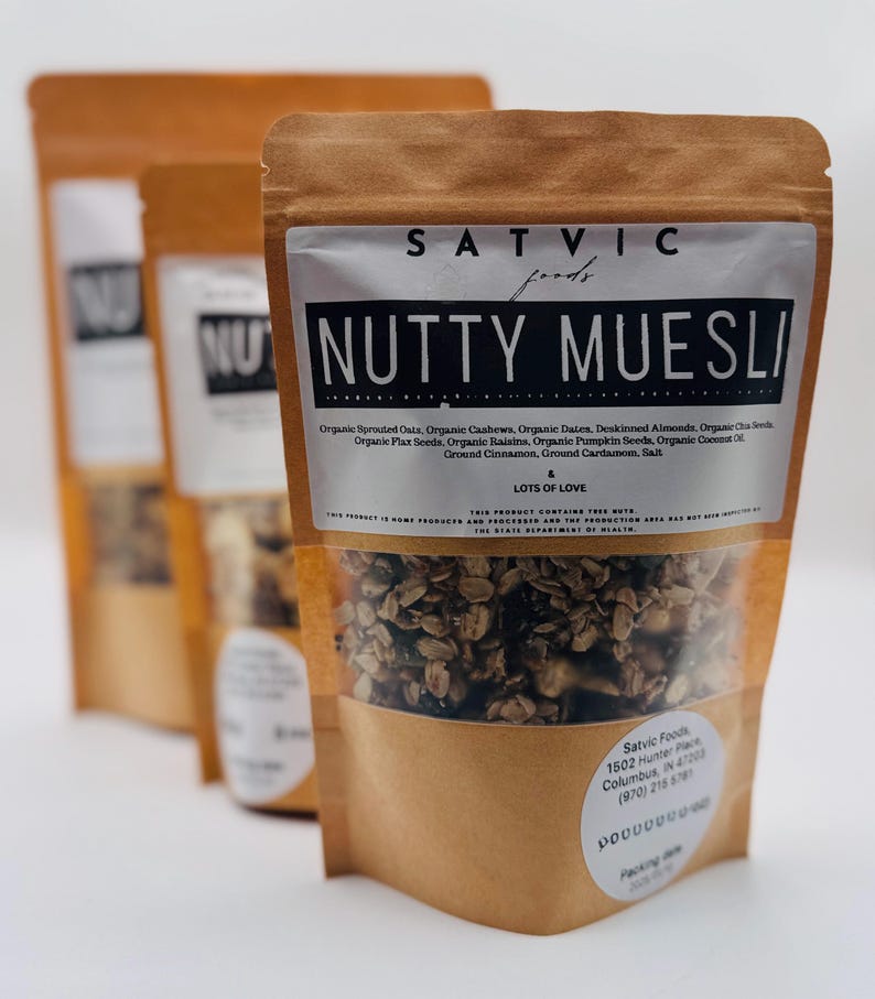 May include: Three brown paper bags of "Nutty Muesli" are displayed. The front bag has a clear window showing the granola inside. The label reads "Satvic Foods" and lists ingredients like oats, cashews, and dates. The text "Lots of Love" is also visible.