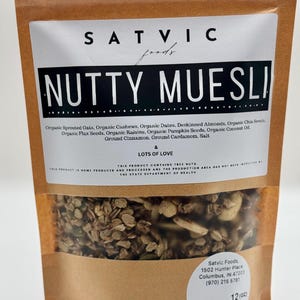 May include: A brown paper bag of "NUTTY MUESLI" from Satvic Foods. The bag features a clear window showing the granola mix of oats, nuts, seeds, and spices. The label lists ingredients and the words "& LOTS OF LOVE."