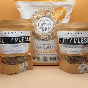 May include: An assortment of organic oat products, including bags of "Nutty Muesli" and "Sprouted Rolled Oats." The packaging features text such as "Gluten Free" and "Lots of Love." The background is a solid orange color.