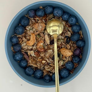 May include: A blue bowl filled with granola, cashews, and blueberries. A gold spoon rests in the center of the bowl. The granola is a mix of grains and nuts. The blueberries are arranged around the edge of the bowl.