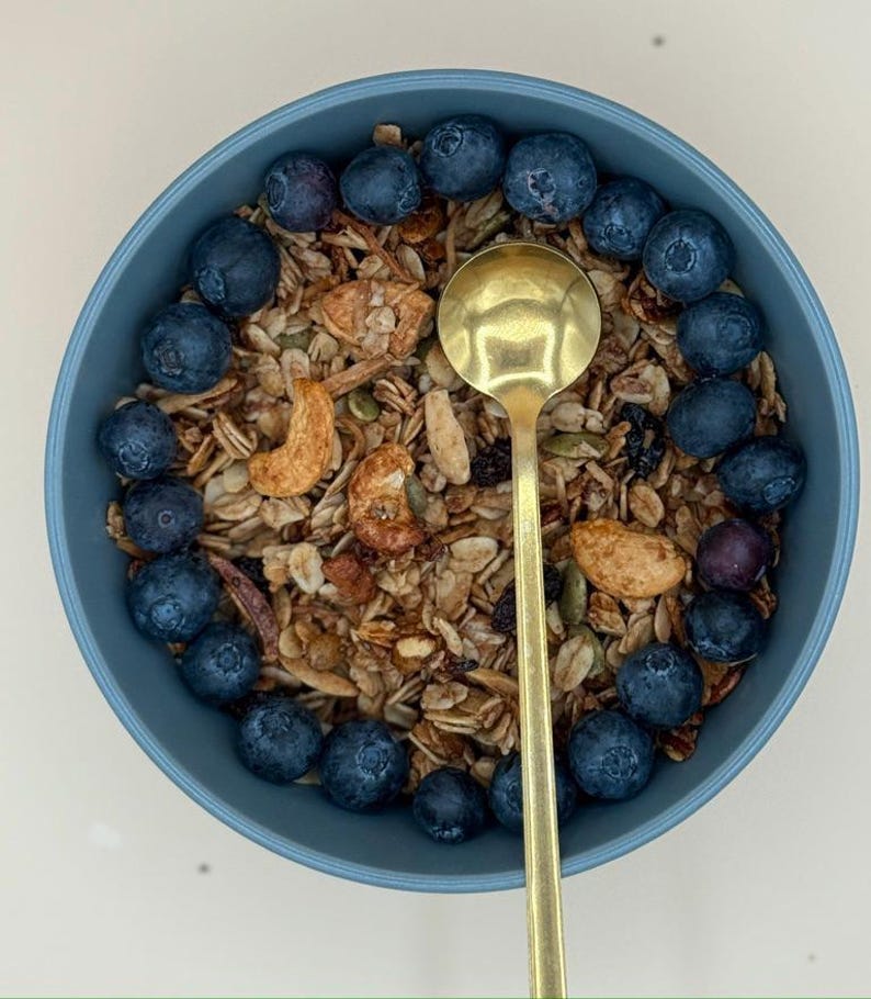 May include: A blue bowl filled with granola, cashews, and blueberries. A gold spoon rests in the center of the bowl. The granola is a mix of grains and nuts. The blueberries are arranged around the edge of the bowl.